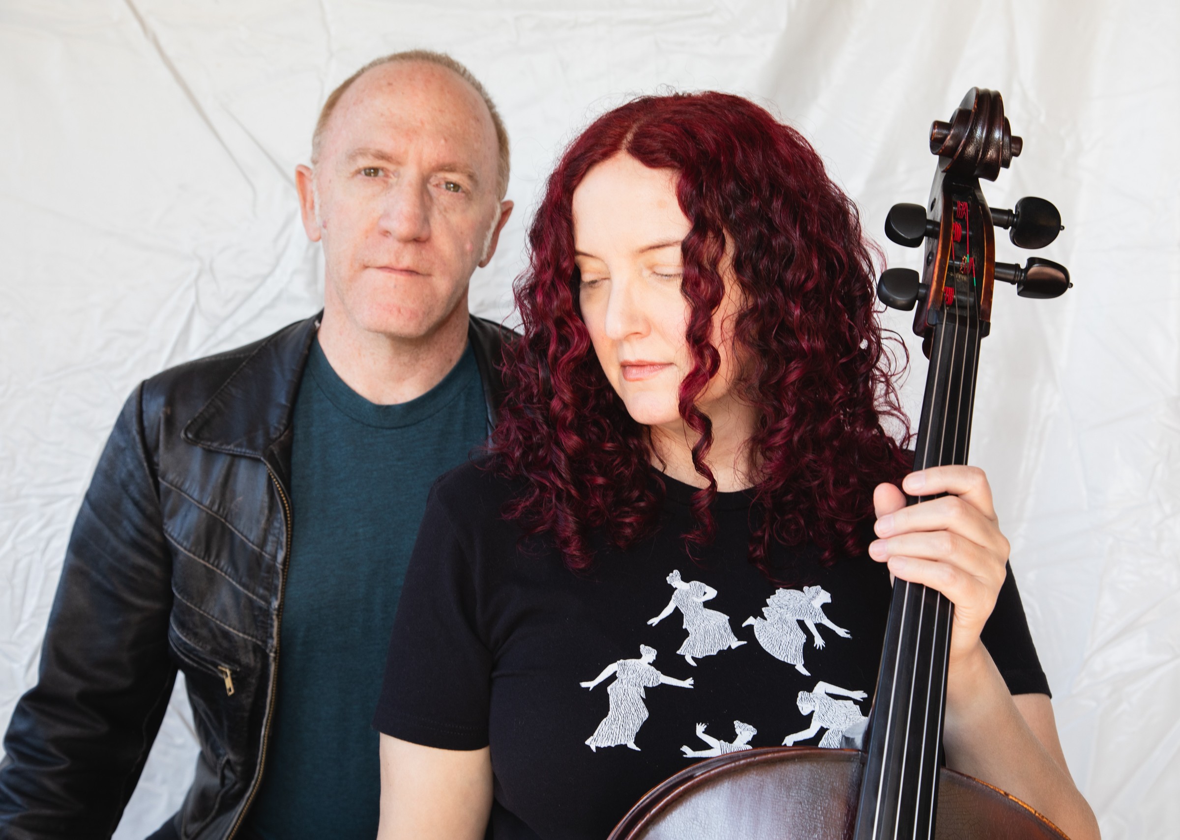 A woman with curly red hair holding a violin, and a man standing behind her, both in front of a white background.