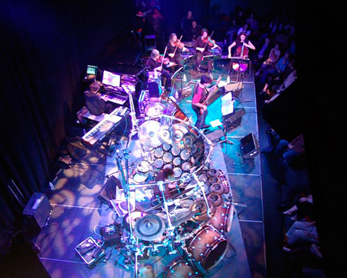 Musicians perform on stage with drums, keyboards, and string instruments, audience seated nearby, stage lit with blue and purple lights.