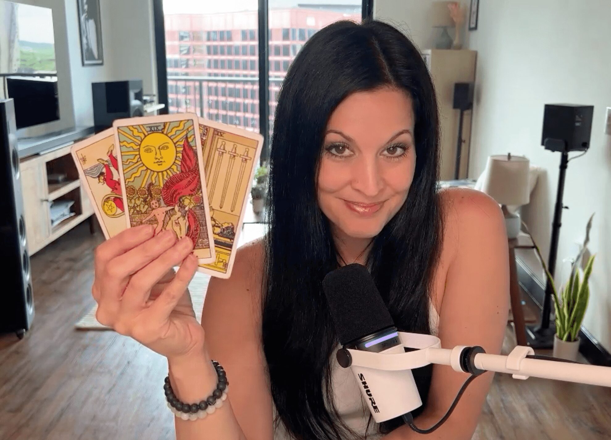 Woman with long dark hair holding tarot cards, sitting in a room with large windows and a microphone.