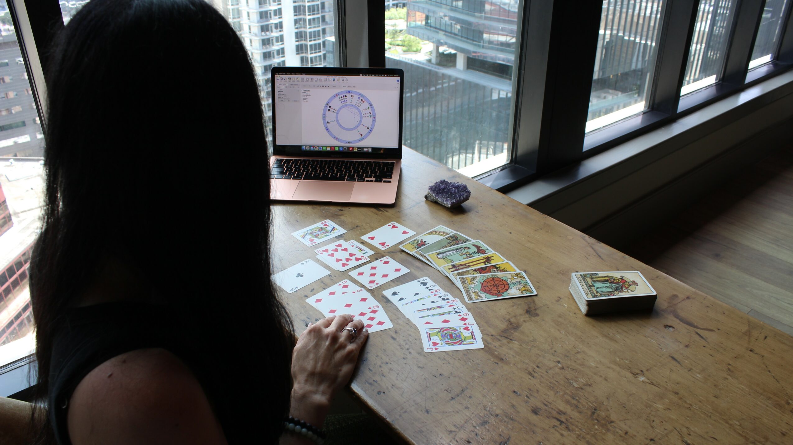 Person playing cards at a wooden table near large windows with city view, laptop, and card deck present.