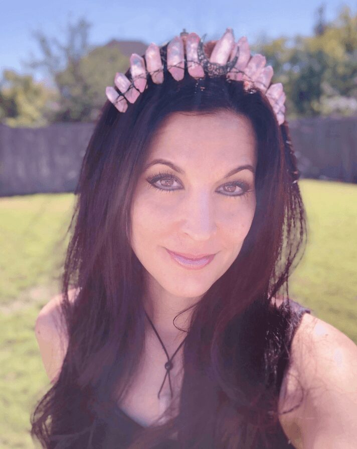 Woman with dark hair wearing a pink crystal crown outdoors, smiling, with trees and a fence in the background.