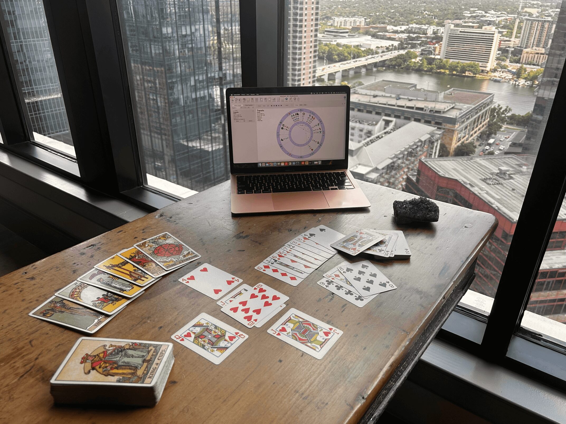 Laptop, playing cards, and papers on a wooden table near a window with a city view.