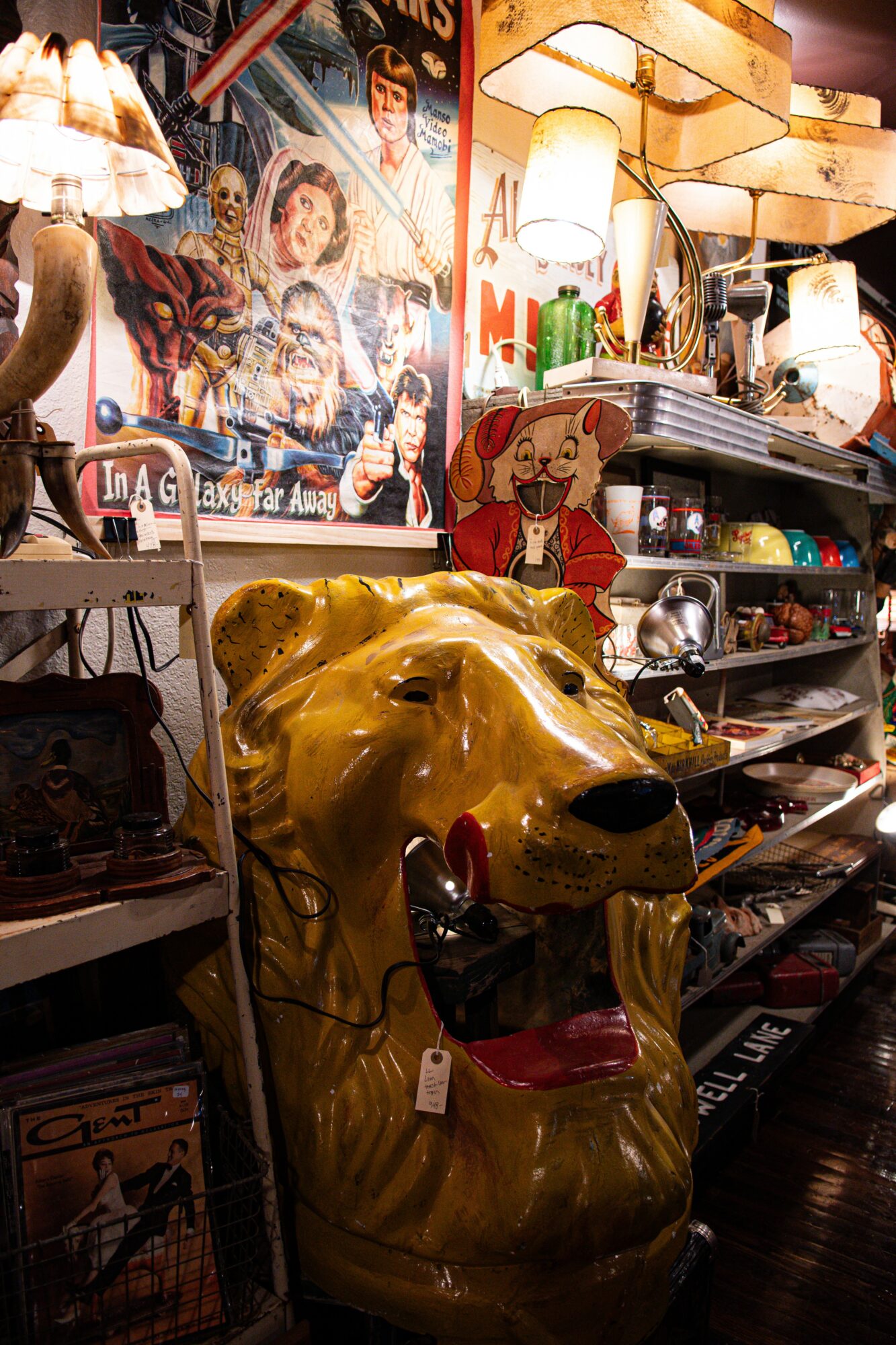 Large yellow ceramic dog statue with a red tongue, surrounded by vintage lamps and collectibles on shelves, colorful posters in background.