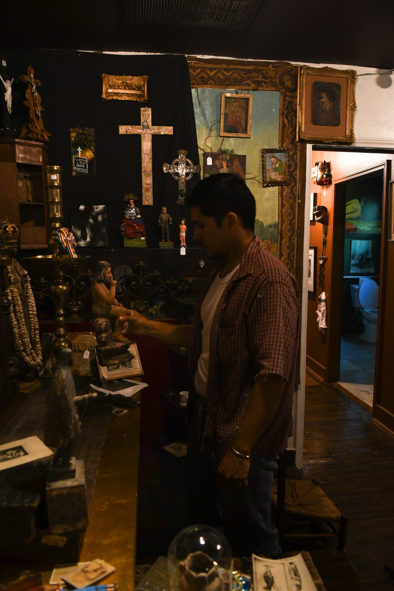 Young man in a checkered shirt browsing religious artifacts in a dimly lit room with crosses and framed pictures.