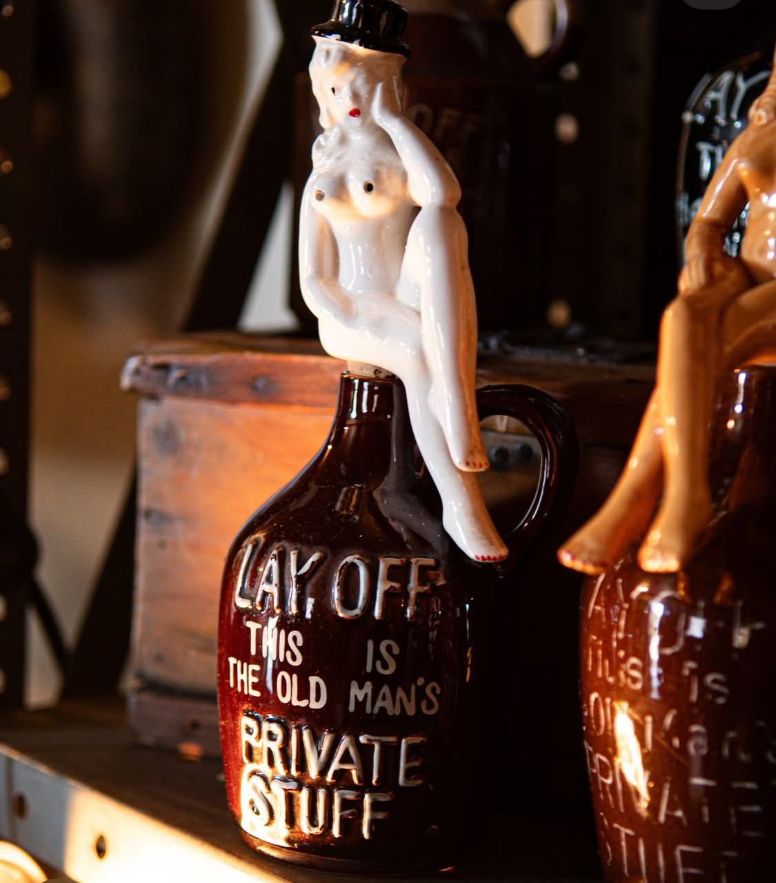 Ceramic figurine of a woman sitting on a bottle with text, placed on a wooden surface, with other objects in background.