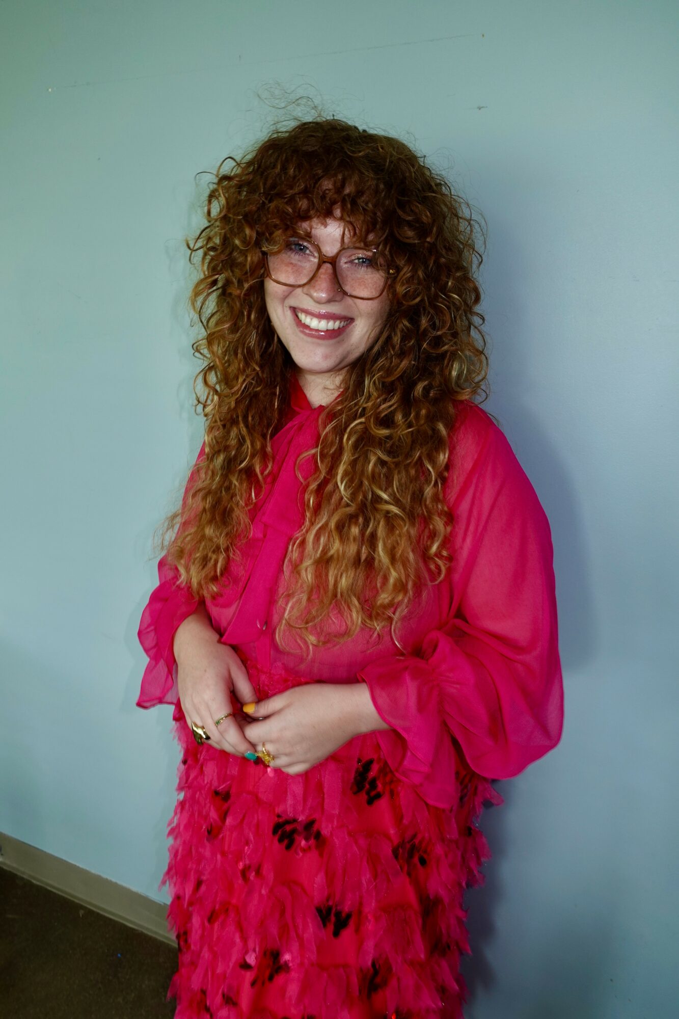 Woman with curly hair and glasses smiling, wearing a bright pink dress with feathered details, standing against a light blue wall.