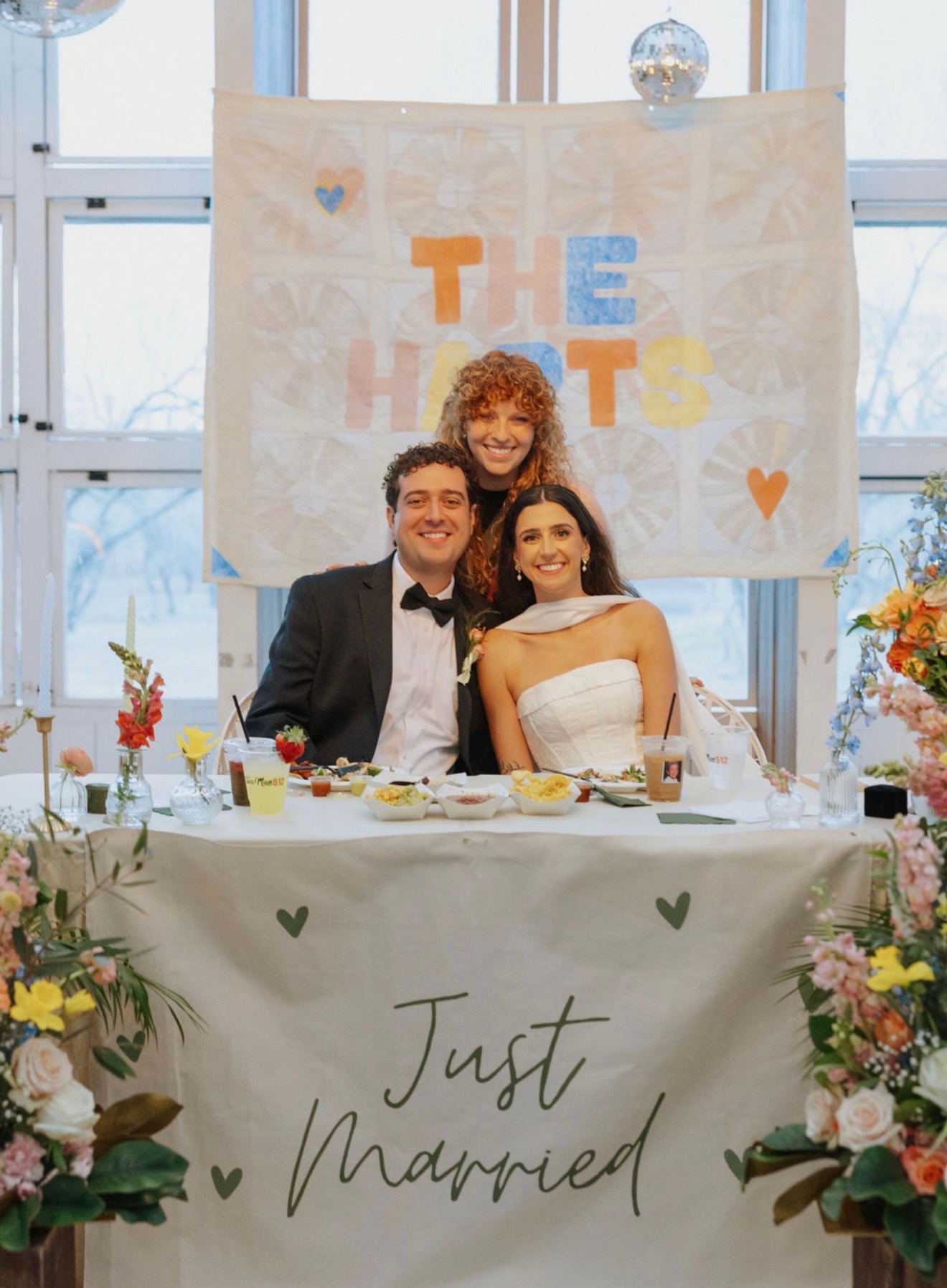 Three people at a wedding reception, two seated and one standing behind, smiling, with a decorated table and a banner in the background.