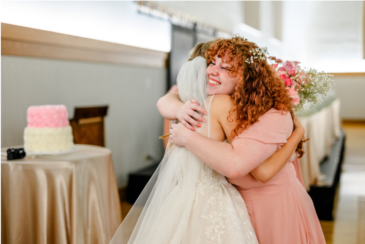Two women hugging, one in a wedding dress, smiling, in a decorated indoor setting with a table and cake in background.
