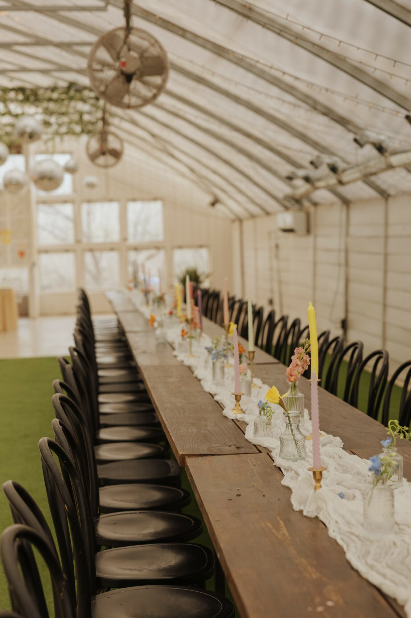 Long table set with candles and flowers inside a tent, with chairs on both sides and hanging lights overhead.