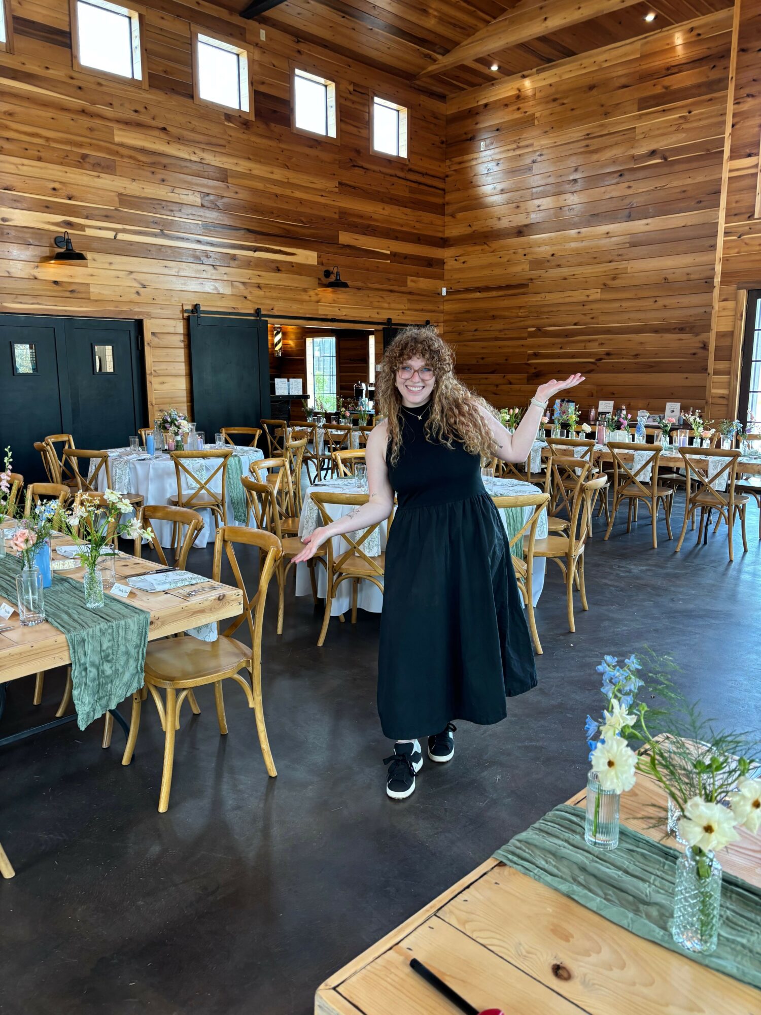 Person with curly hair wearing a black dress and sneakers, standing in a wooden room with tables and chairs, reaching out.