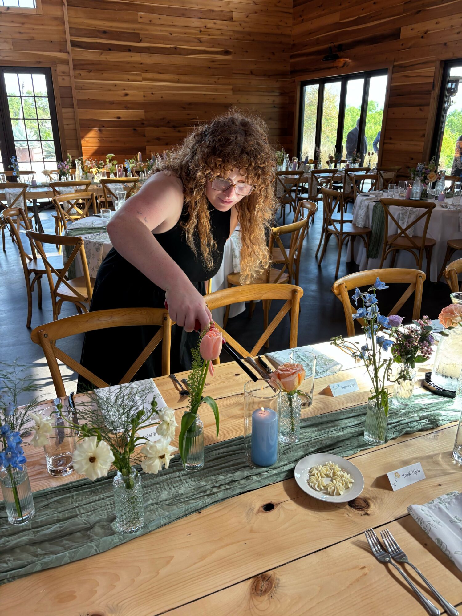 Young girl with curly hair arranging flowers on a table in a cozy wooden room, with chairs and windows in the background.