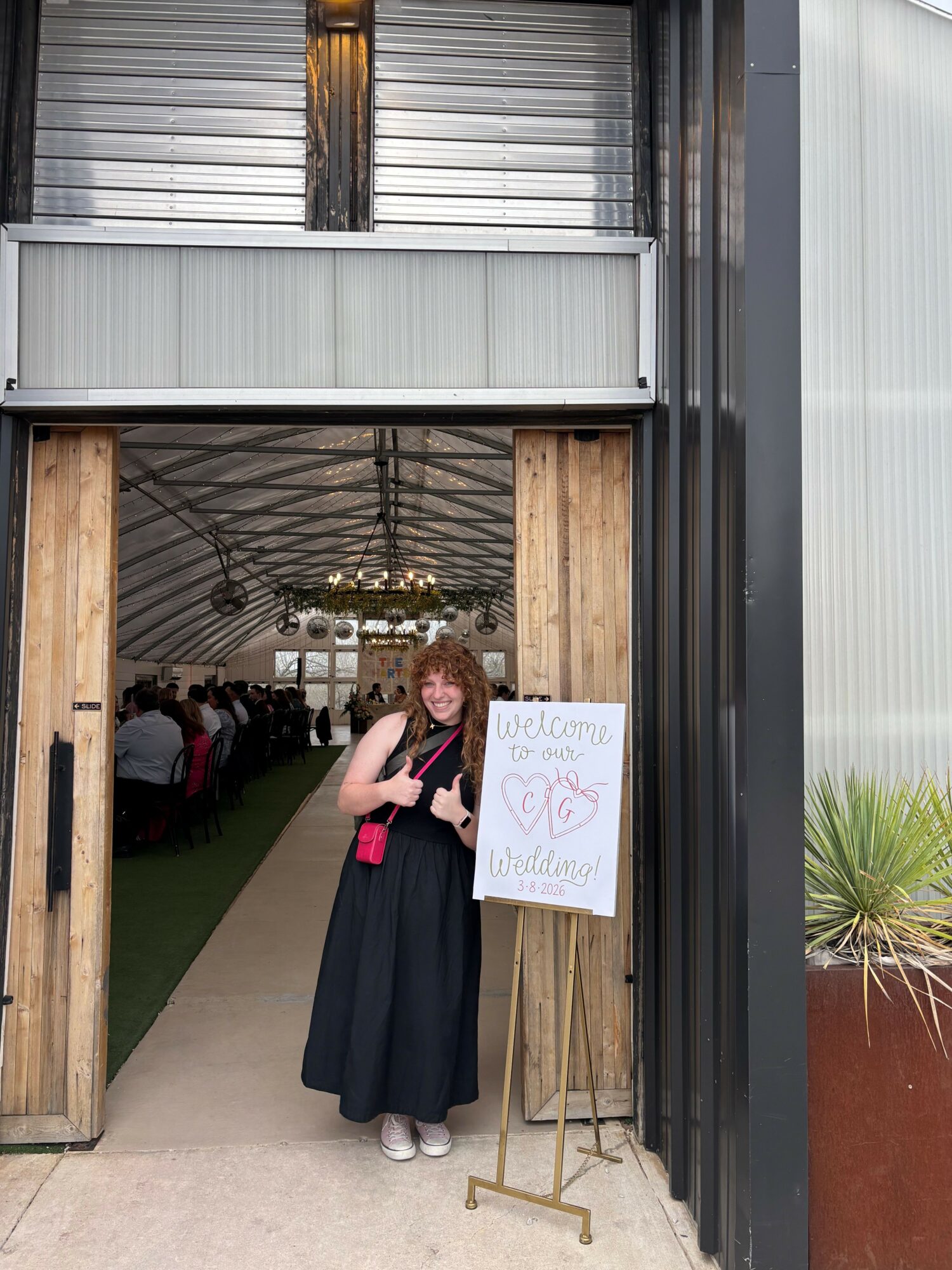 Woman in black dress standing outside near a wooden structure, holding a pink item, with a signboard nearby.
