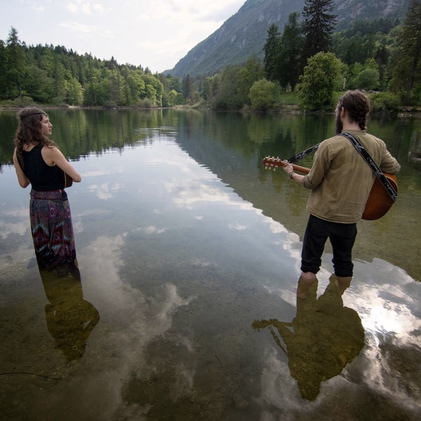 Two people stand in a river with mountains and trees in the background, one playing guitar, the other watching.