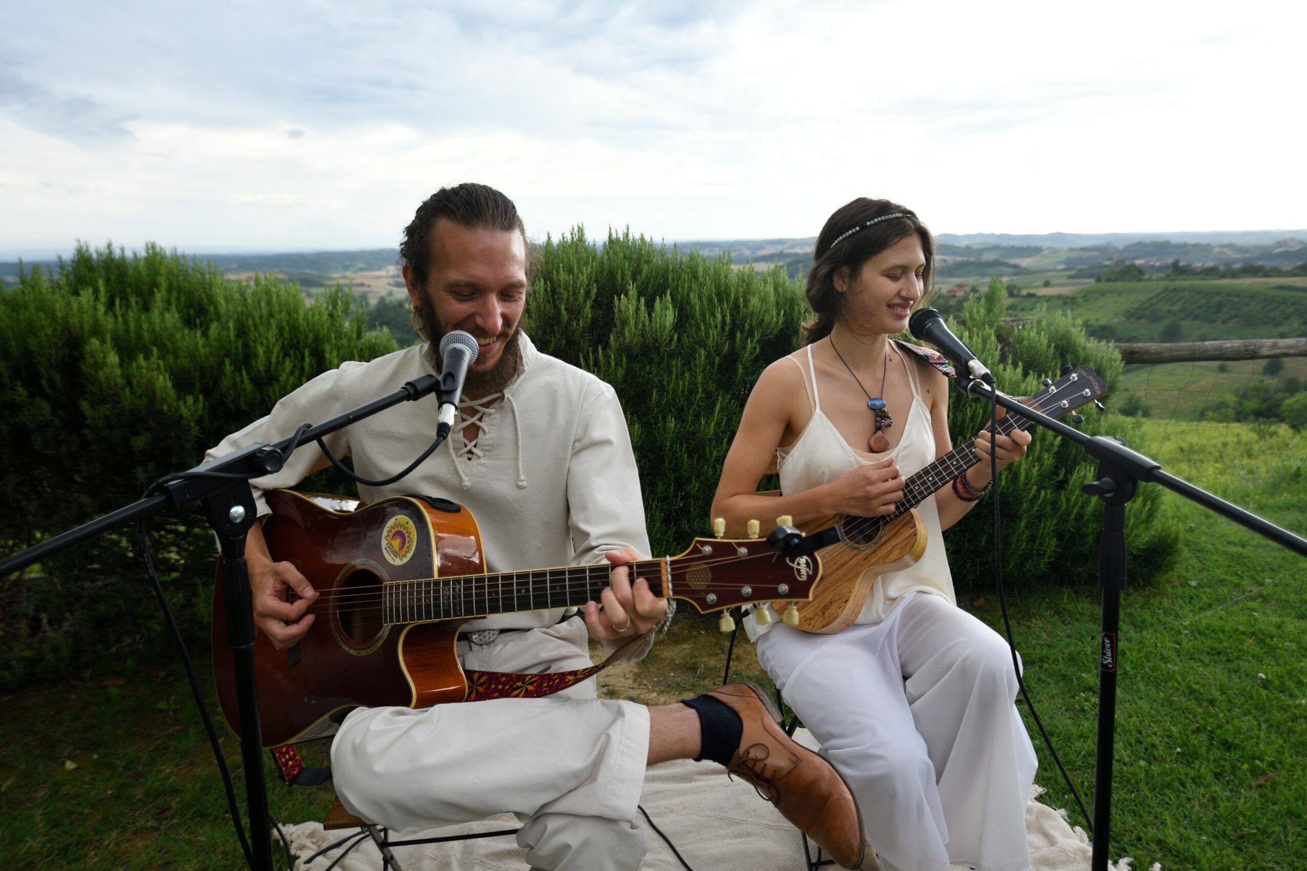 Two people playing guitars and singing outdoors with green fields and bushes in the background.