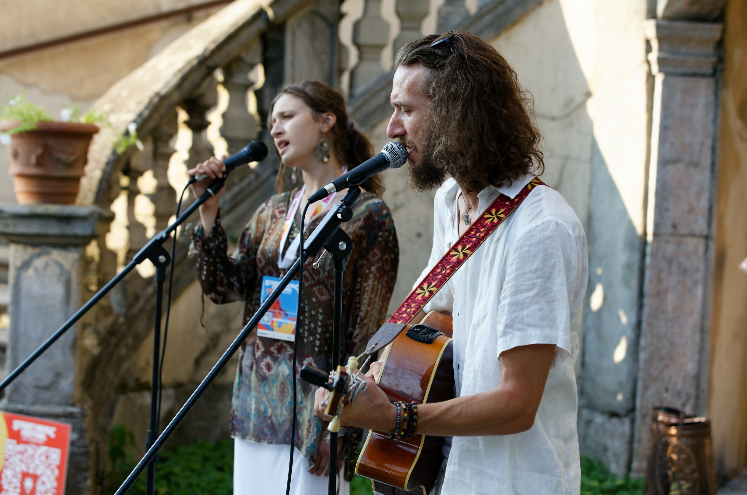 Two people perform music outdoors, one singing into a microphone and the other playing guitar, with stone stairs in background.