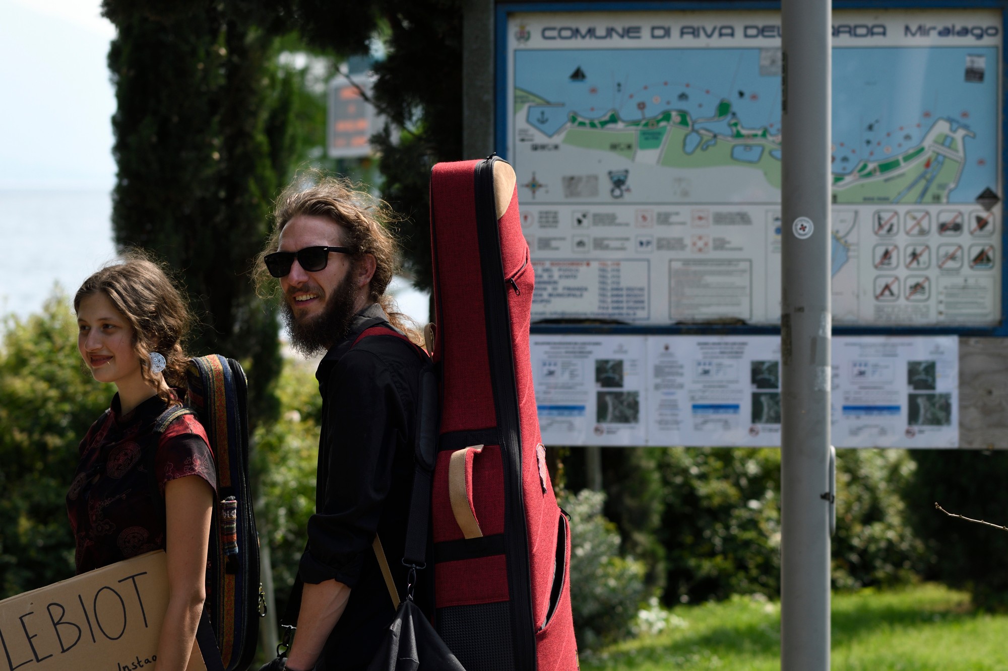 Two people with backpacks walking outdoors near a park information board, smiling and wearing sunglasses.