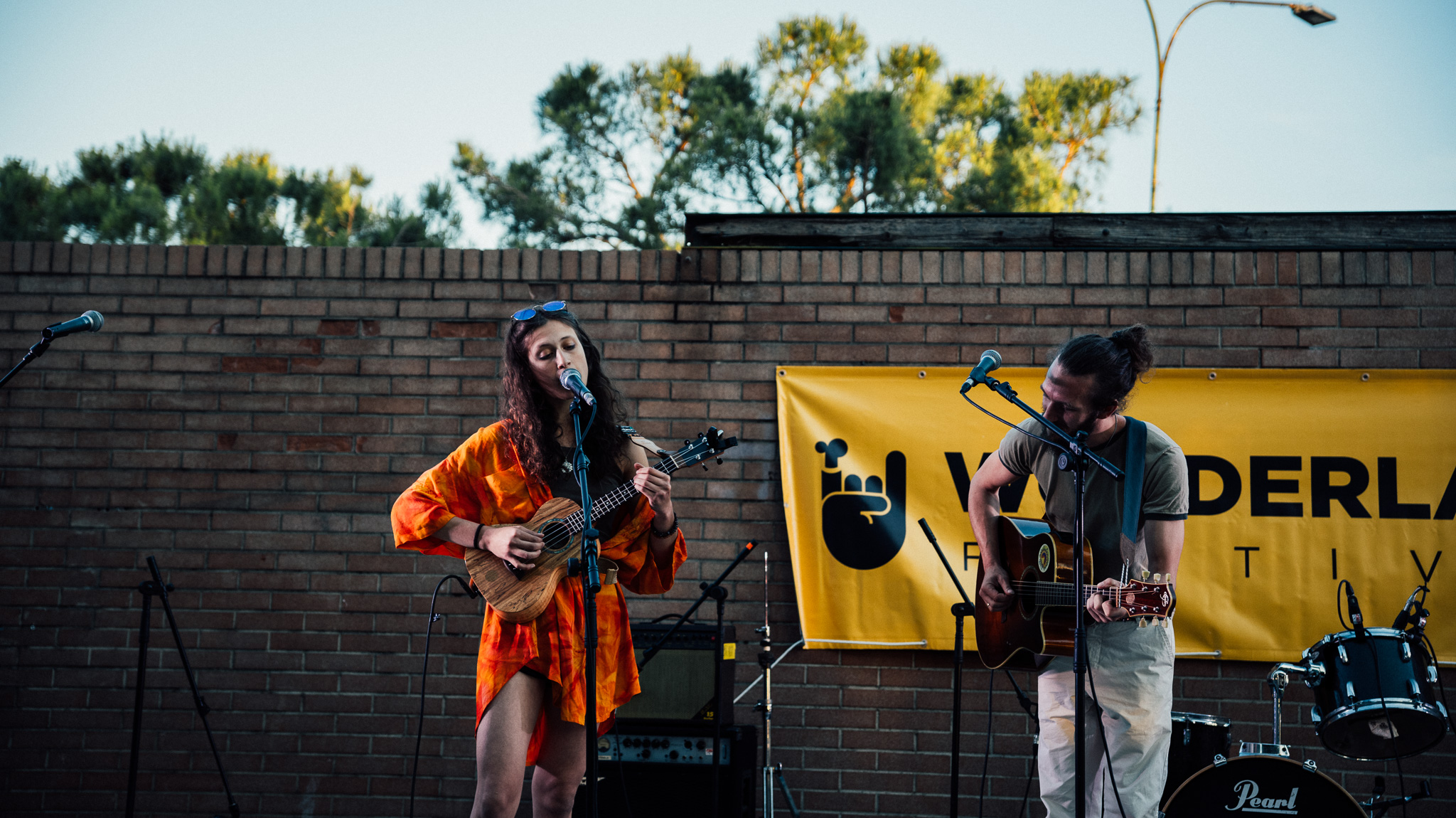 Two women perform with guitars outdoors against a brick wall and yellow banner, trees, and sky in background.
