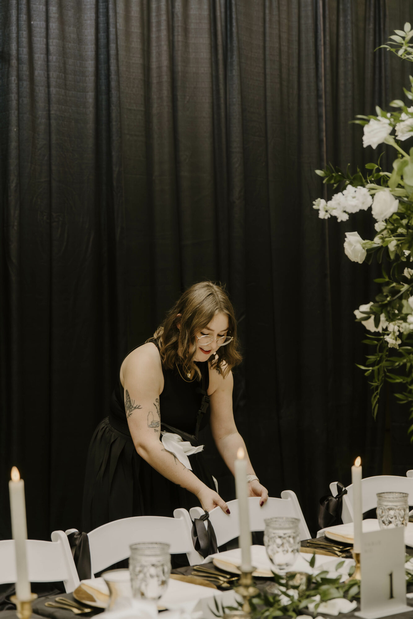 Woman with glasses and tattoos arranging table settings in front of black curtains, with white flowers on the right.