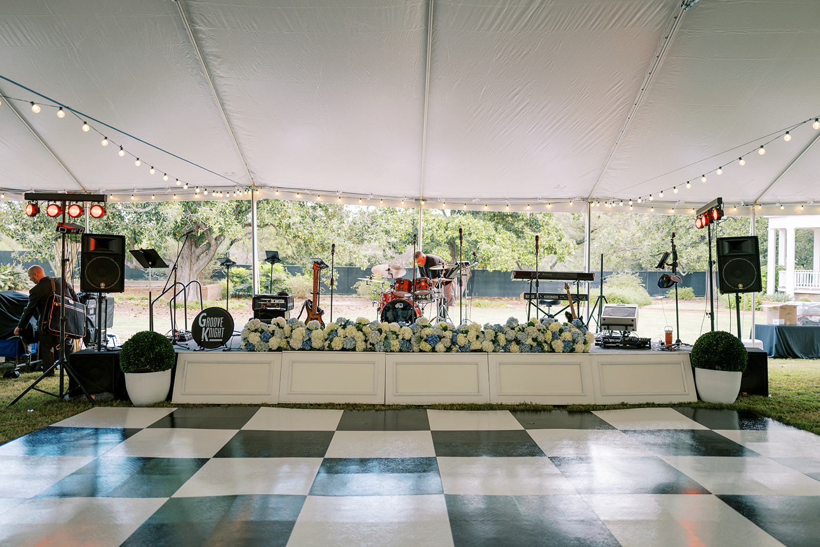 Stage with musical instruments and speakers under a tent, decorated with string lights and potted plants, on a checkered dance floor.
