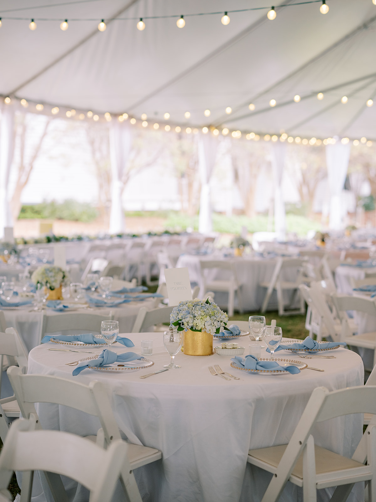 Wedding reception under a tent with decorated tables, string lights, and white chairs, outdoors with trees in the background.