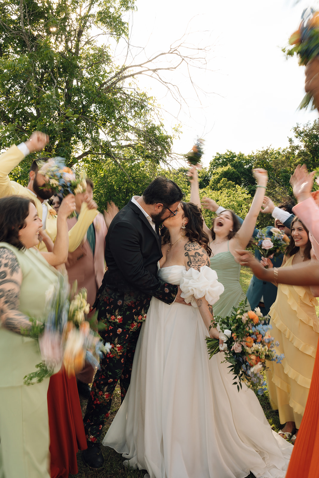 Couple kissing surrounded by friends holding bouquets at outdoor wedding, trees in background.