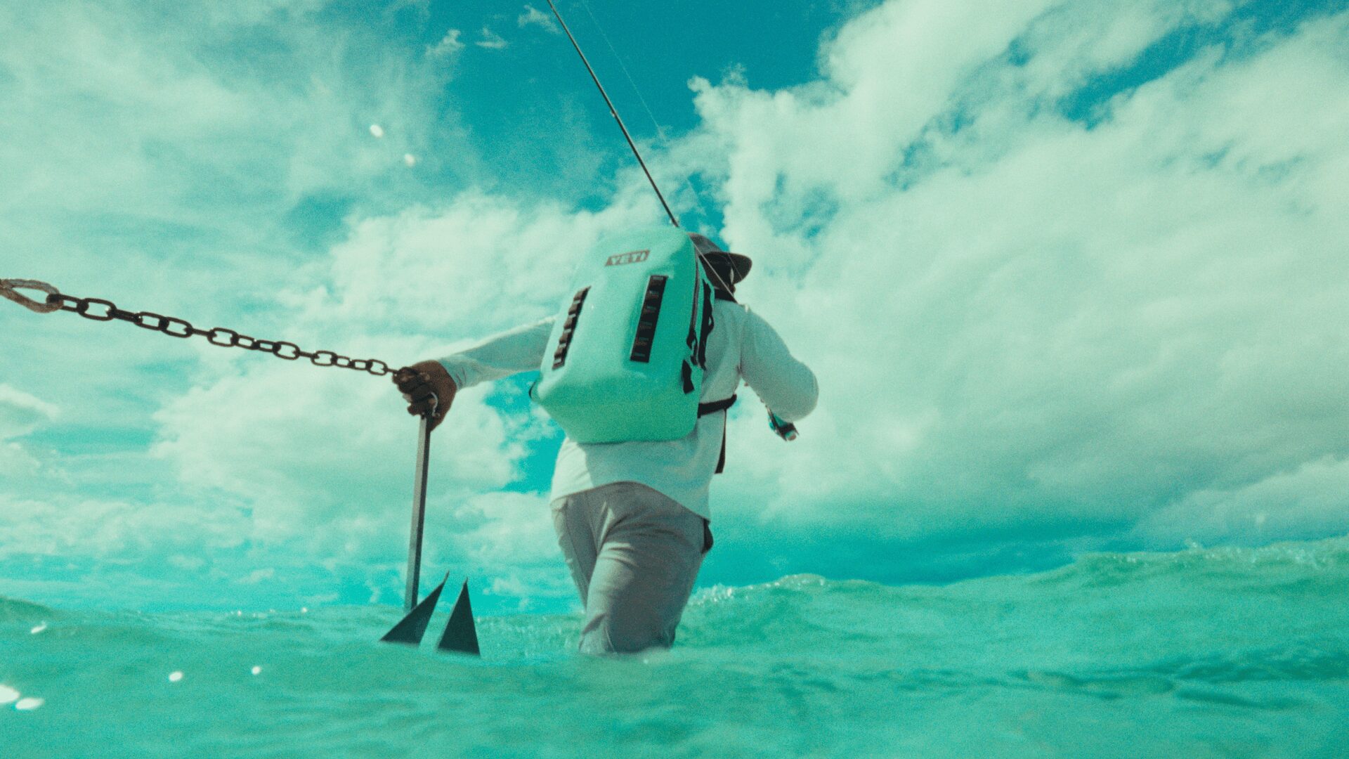 Person in a wetsuit and life vest walking in water holding a pole, with a cloudy sky above.