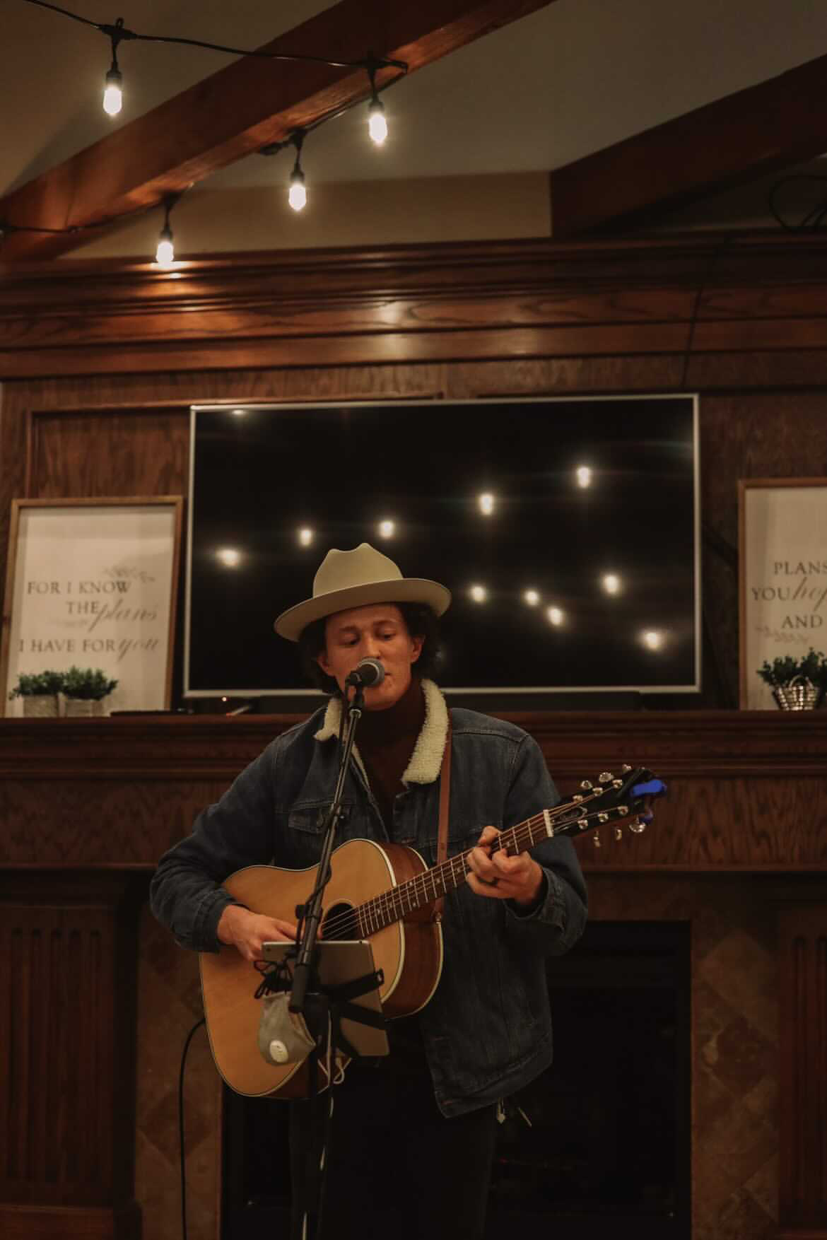 Person wearing a hat playing guitar and singing into a microphone indoors, with warm lighting and wooden decor.