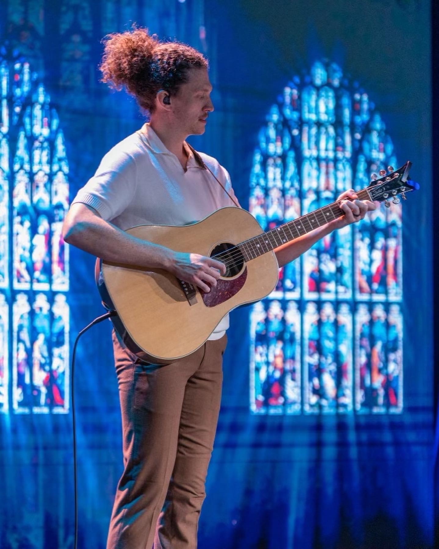 Woman with curly hair playing an acoustic guitar on stage with stained glass windows in the background.