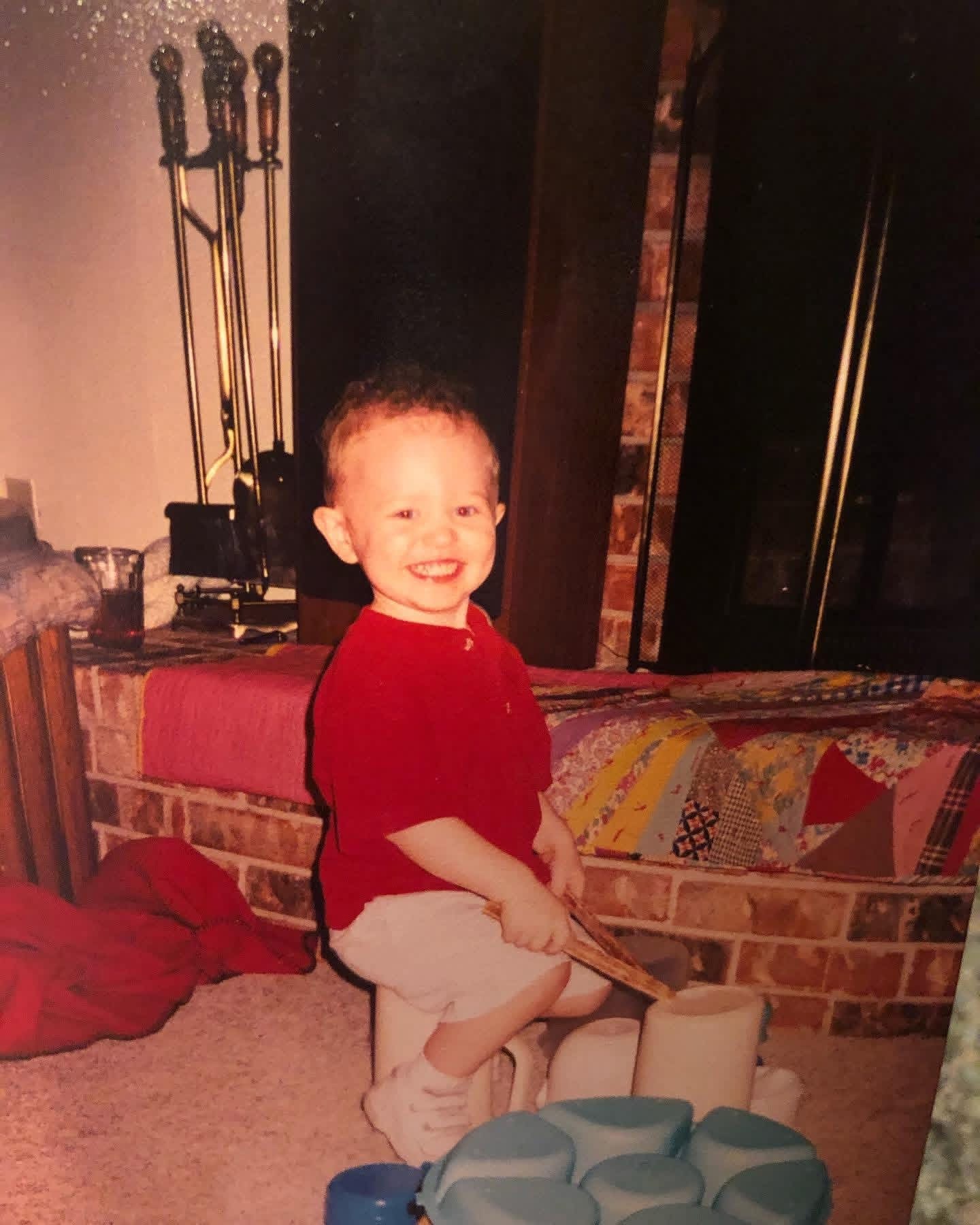 Child with short hair smiling, wearing a red shirt and light-colored shorts, sitting on a stool in a cozy room.