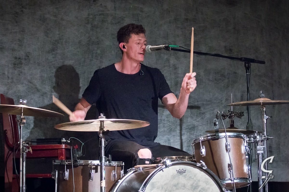 Young man playing drums and singing into a microphone, holding drumsticks, against a plain background.