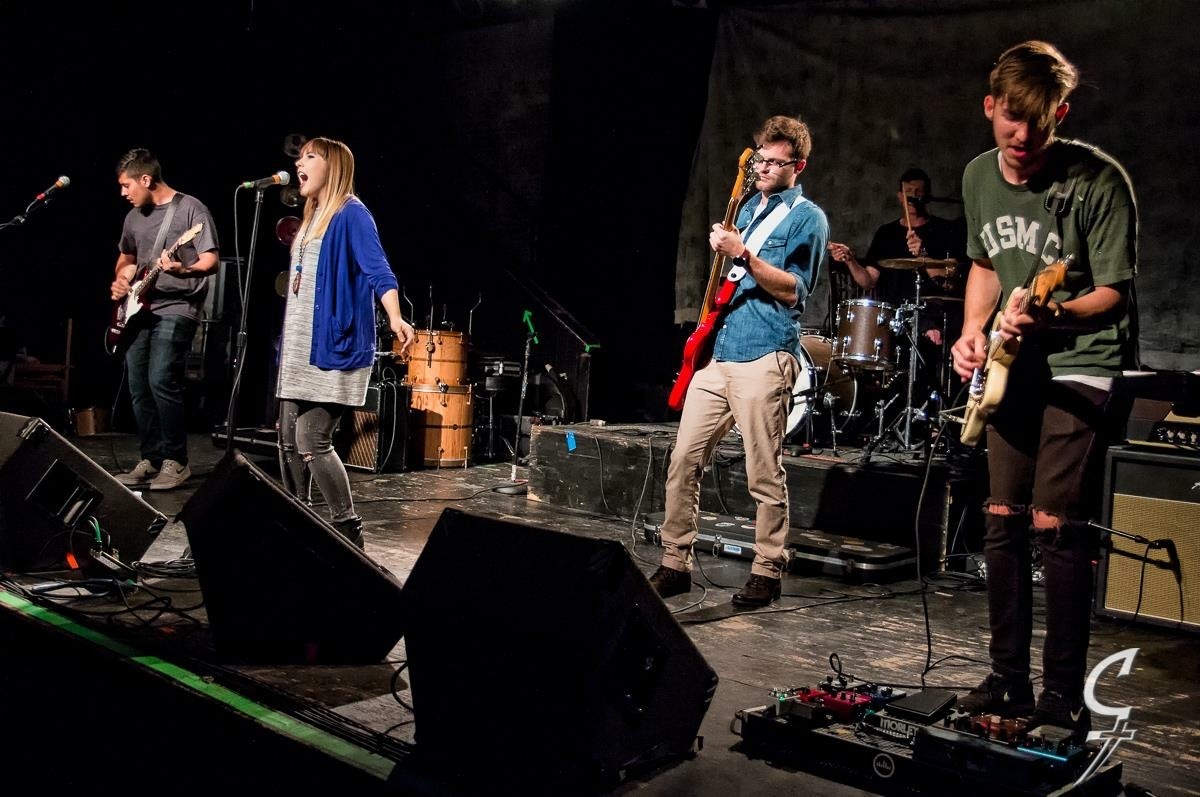 Band performing on stage with four musicians, including guitarists and a drummer, in a dark setting.