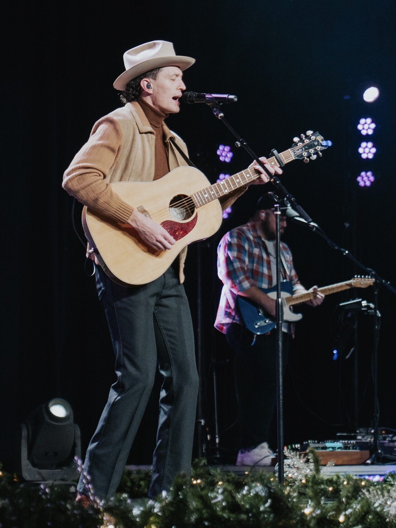 Man wearing a hat playing an acoustic guitar on stage with a person in the background playing electric guitar, stage lights shining.