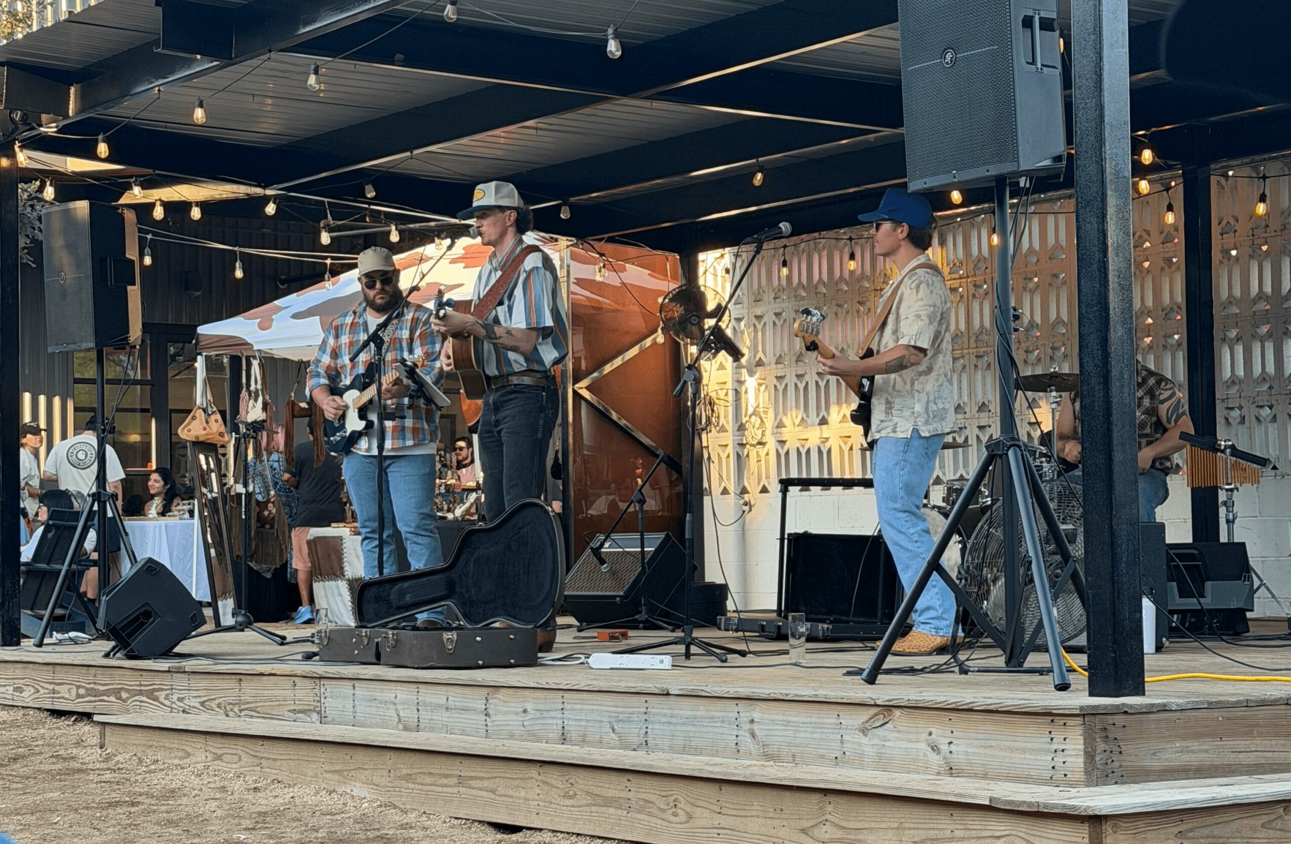 Three musicians perform on an outdoor stage with string lights, instruments, and equipment, under a black canopy.