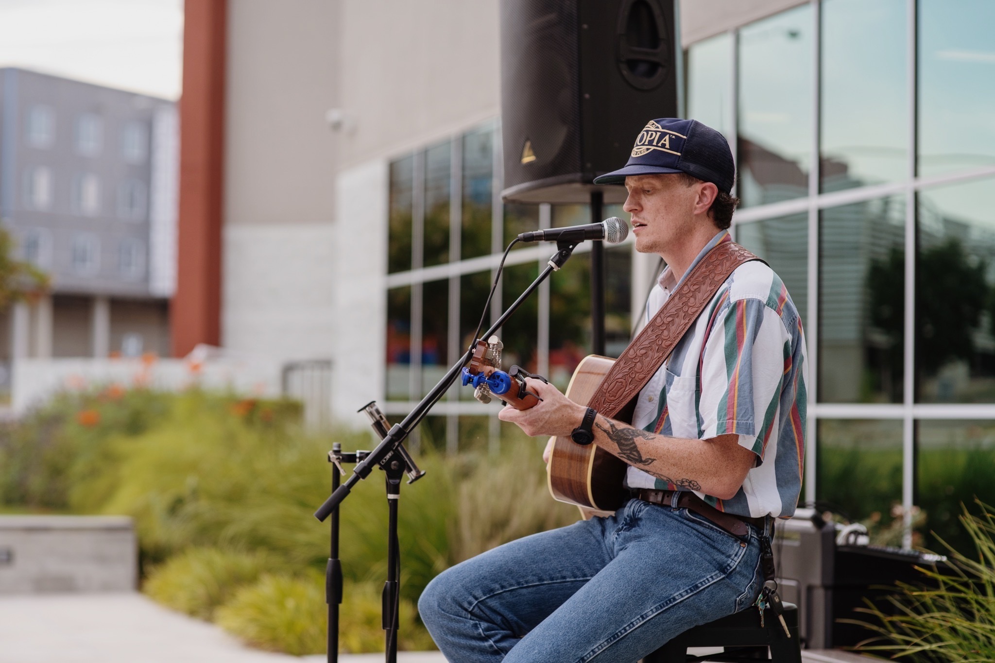 Person playing guitar and singing into a microphone outdoors, with a building and greenery in the background.