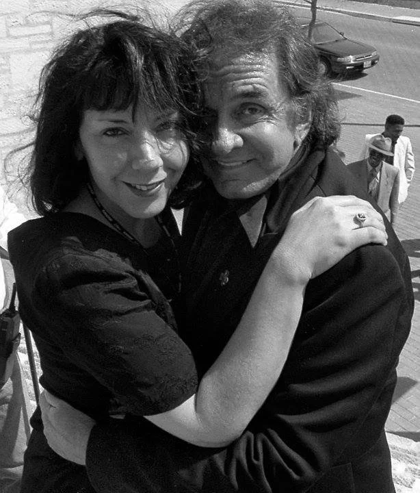 Two people hugging outdoors, smiling, with a street and pedestrians in the background.