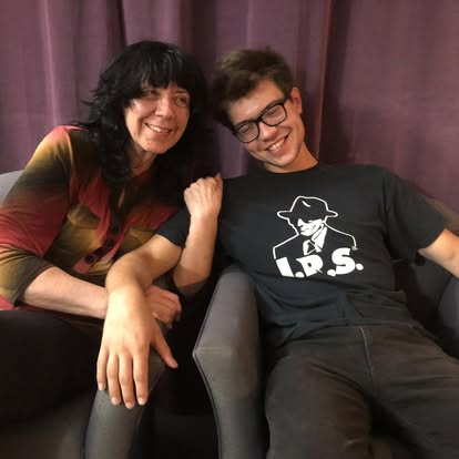 Two people sitting on a couch, smiling, with purple curtains in the background.