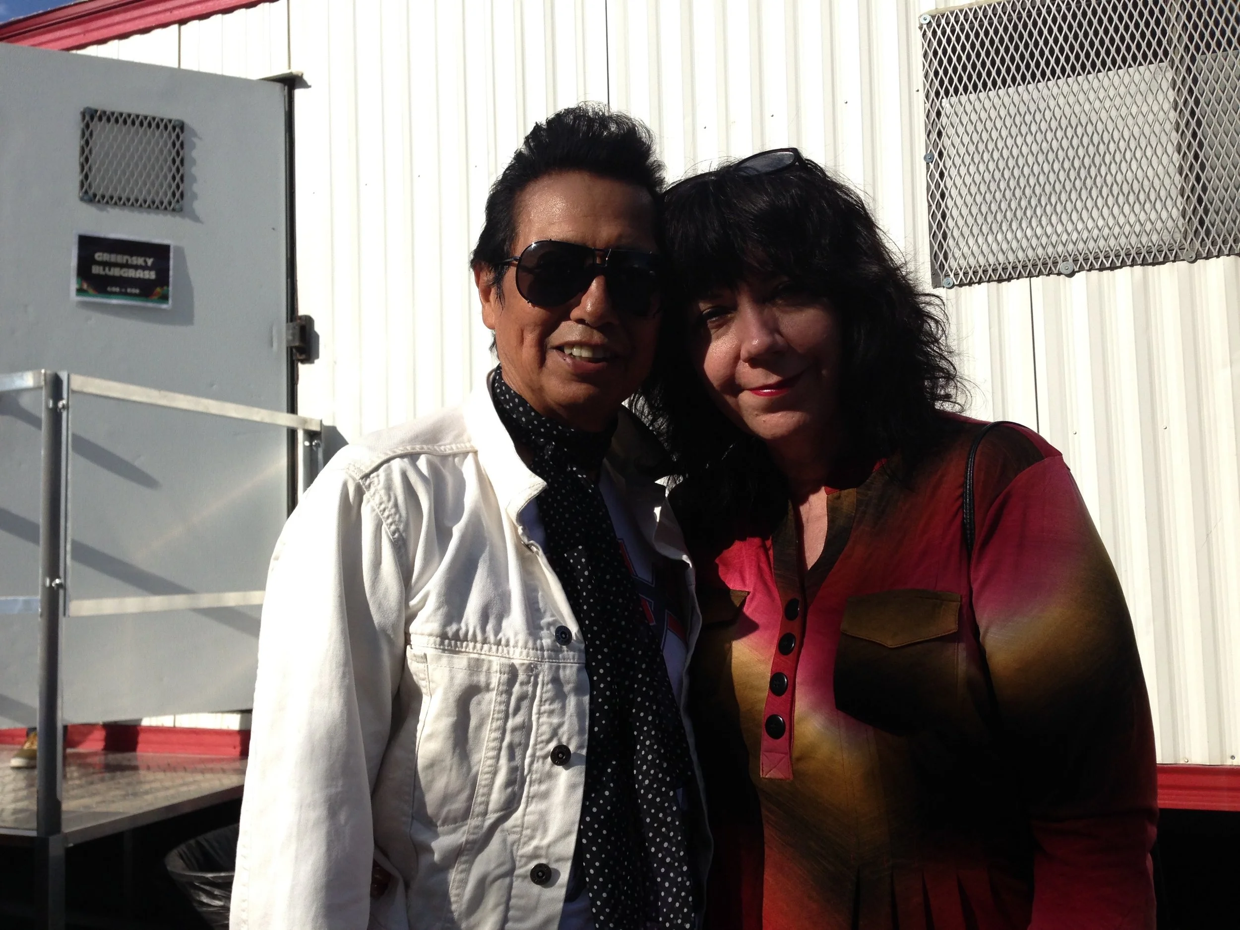 Two people standing close together outdoors, smiling, with a white building and metal structure in the background.