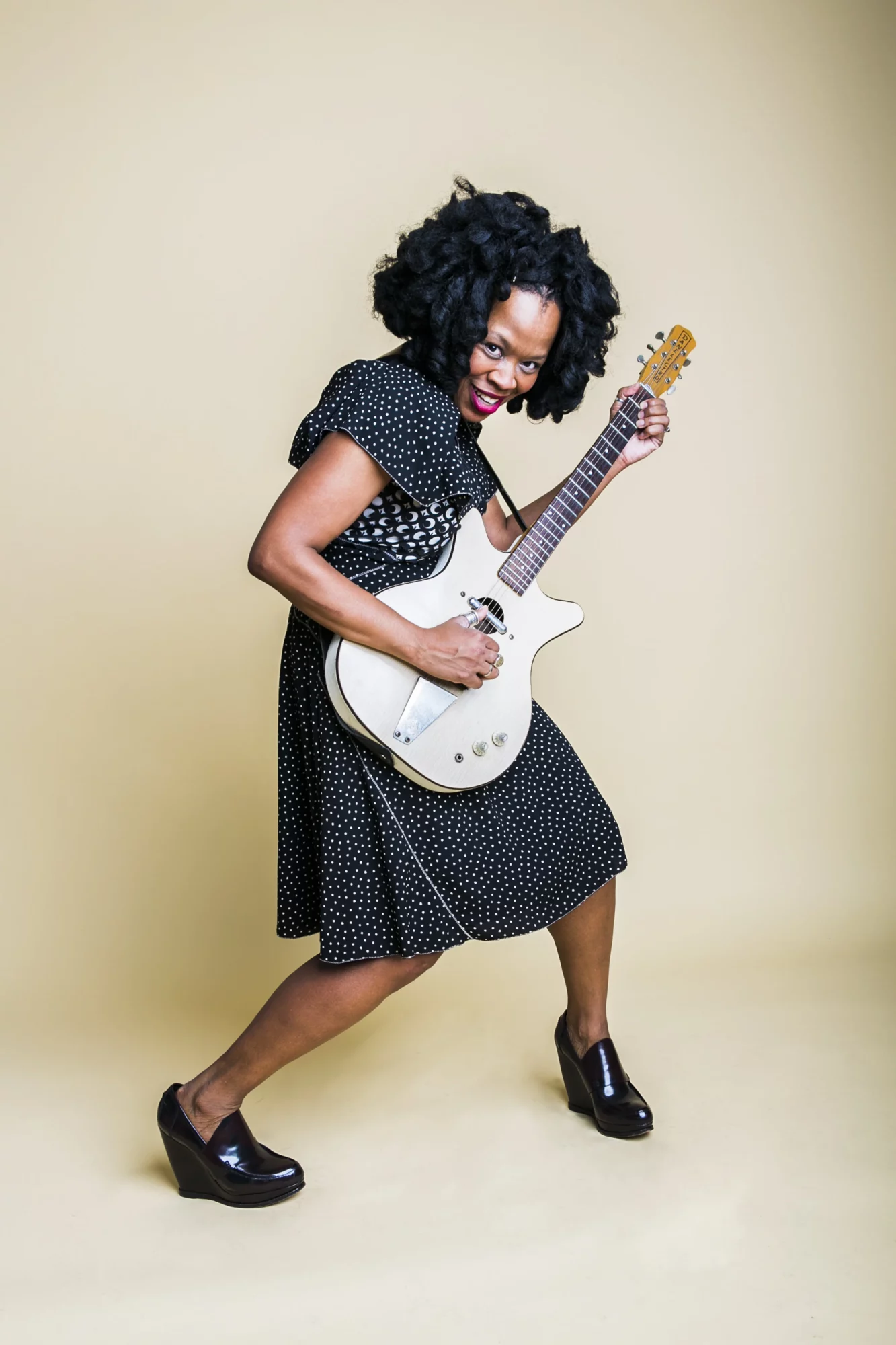 Woman with curly hair playing an electric guitar, wearing a black polka dot dress and high heels, smiling energetically.