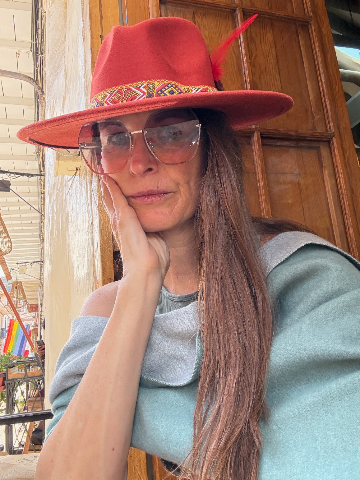 Woman wearing a red hat with a feather, glasses, and a gray top, resting her face on her hand.