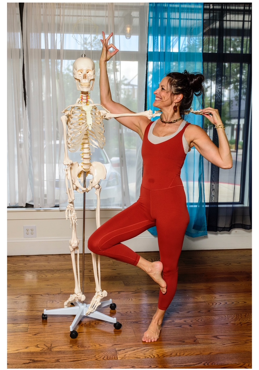 Woman in red jumpsuit practicing yoga next to skeleton model indoors, wooden floor, colorful curtains, smiling, balancing on one foot.