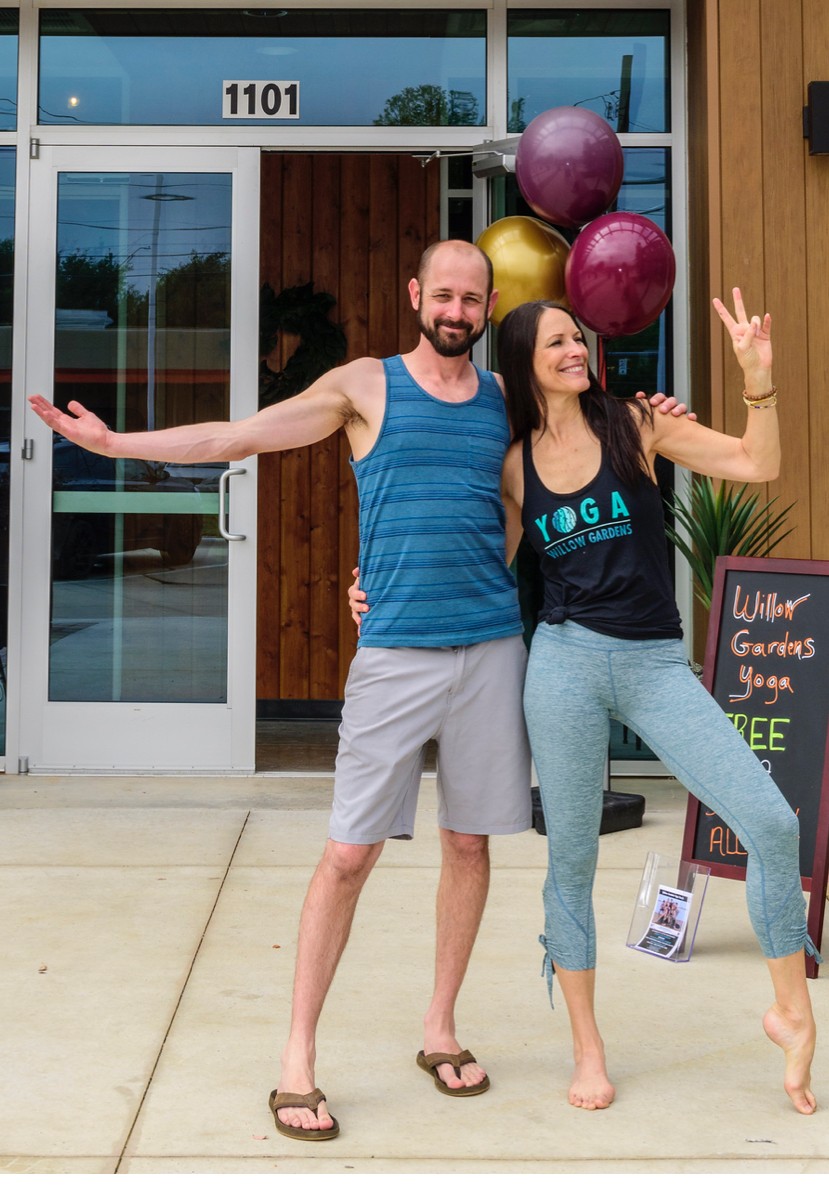 A man and woman stand outside a yoga studio, smiling and making peace signs, with balloons and a chalkboard sign nearby.