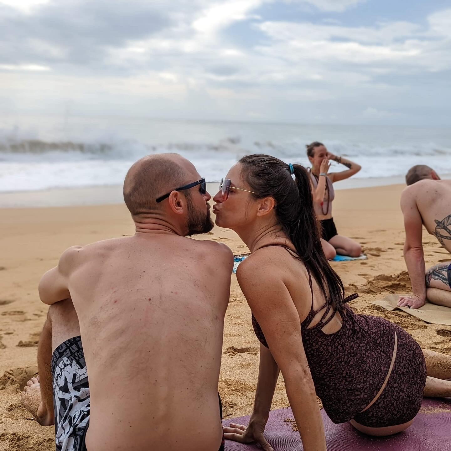 A couple sitting on the beach facing each other, with other people in the background, ocean waves, and cloudy sky.
