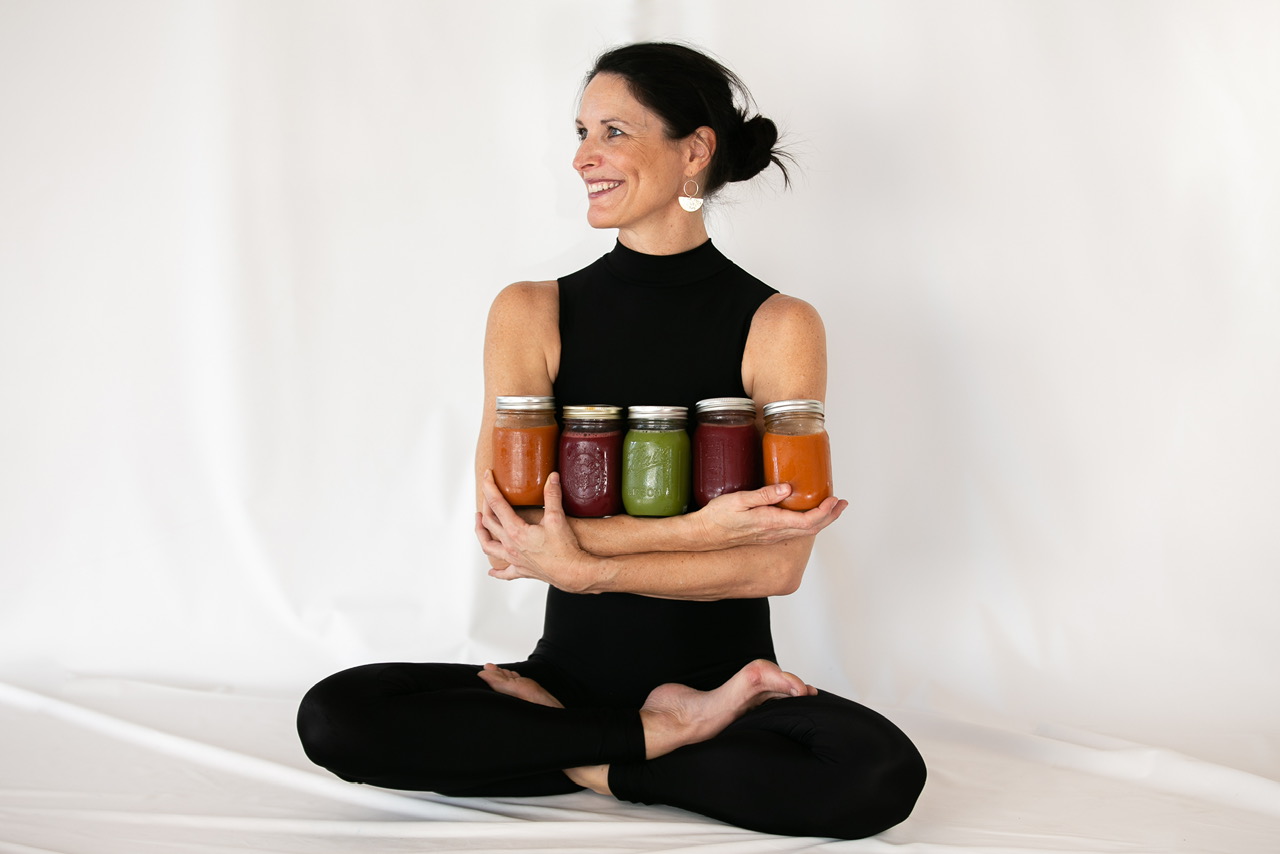 Woman sitting cross-legged holding six jars of colorful smoothies, smiling, against a plain white background.
