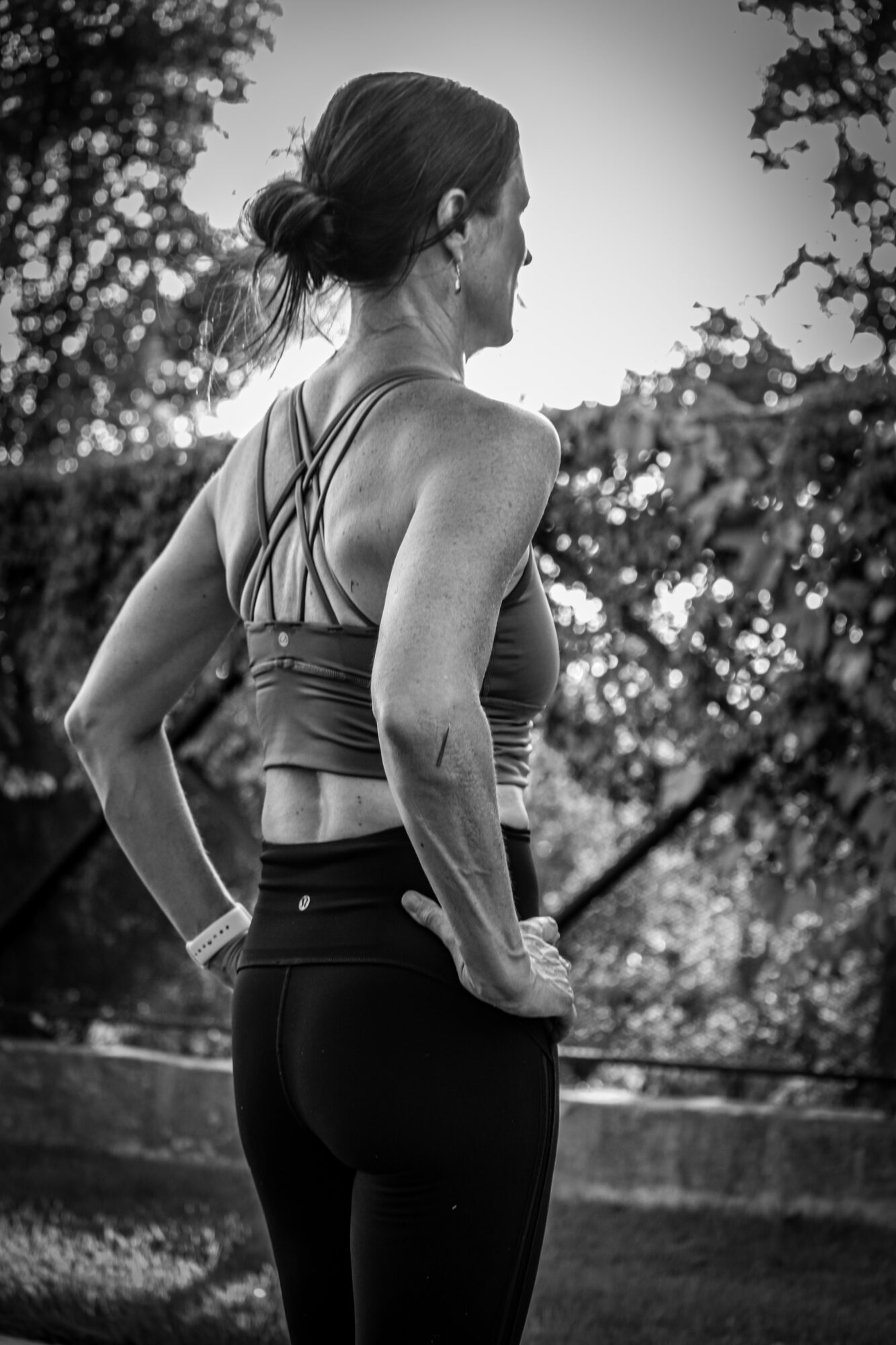 Side view of a woman in athletic clothing outdoors, with trees and sunlight in the background.