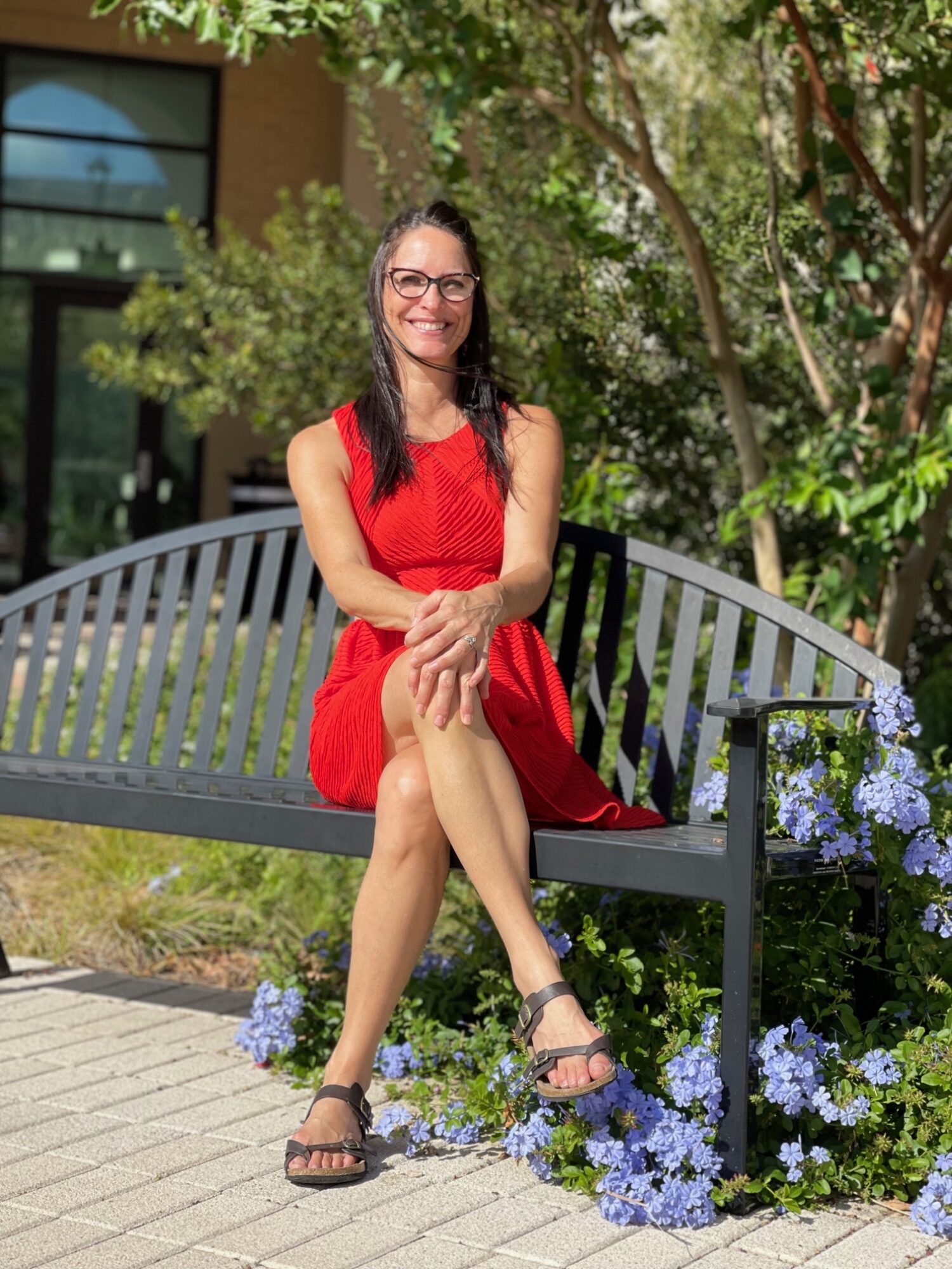 Woman in red dress sitting on a bench outdoors, surrounded by greenery and purple flowers.