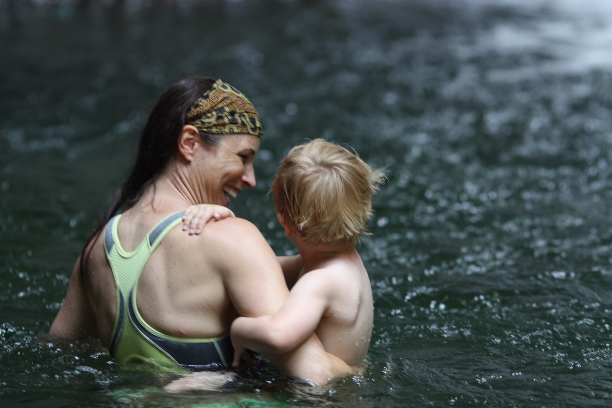 Woman holding a young child in water, both smiling, outdoors with water background.