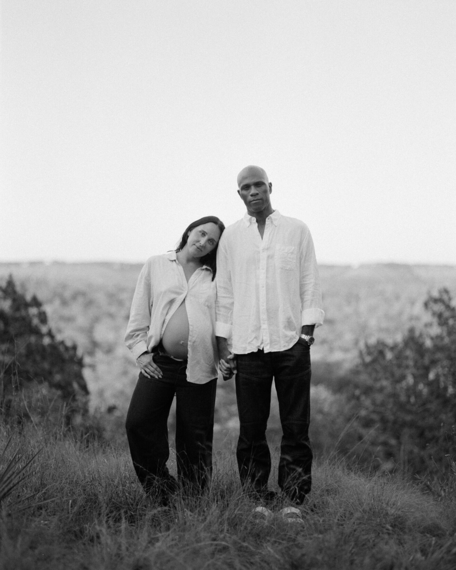 A man and woman stand outdoors on grass, with a landscape and sky in the background, both looking at the camera.