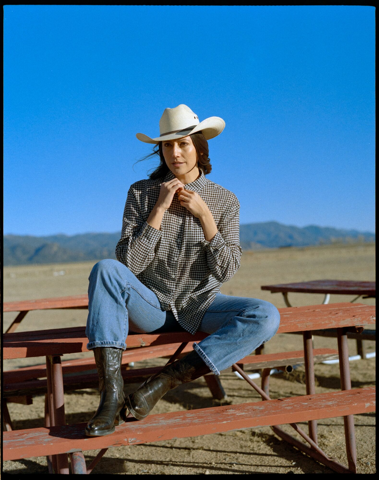 Woman wearing a cowboy hat, sitting on a picnic table outdoors with mountains in background, blue sky, casual clothing, and boots.