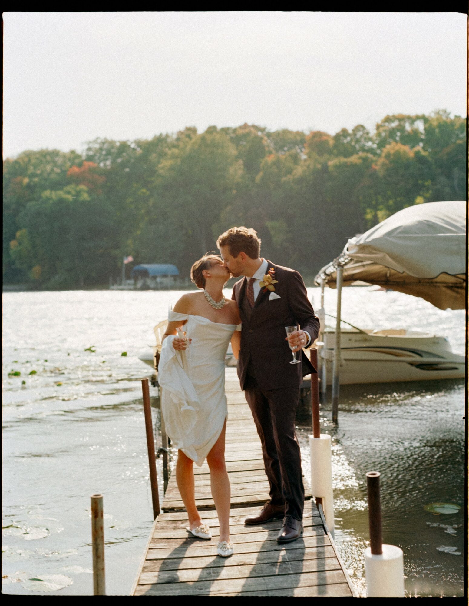 A couple in wedding attire standing on a dock by a lake, with trees and boats in the background.