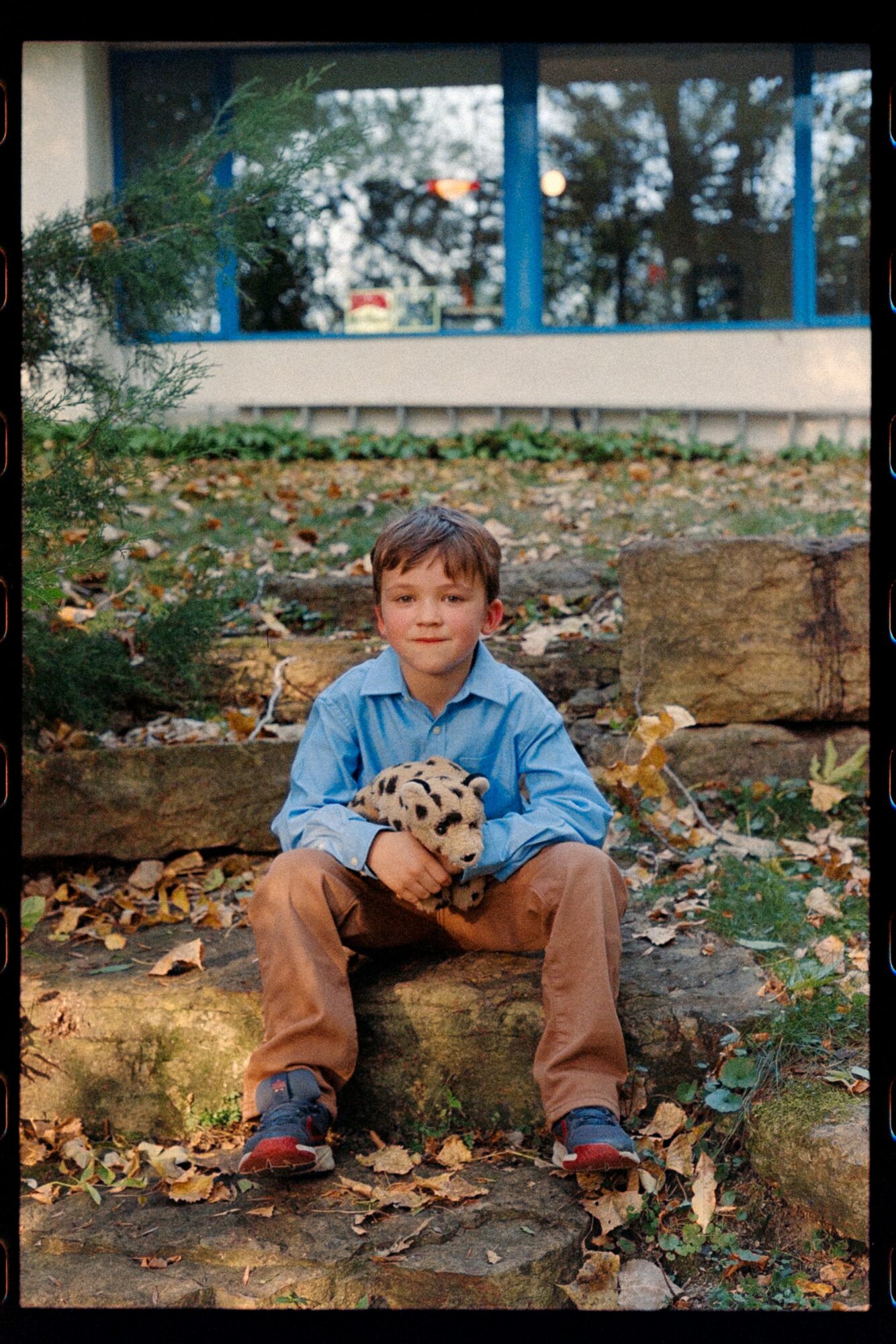 Young boy sitting on rocks outdoors holding a stuffed animal, surrounded by fallen leaves and trees, with a building in the background.